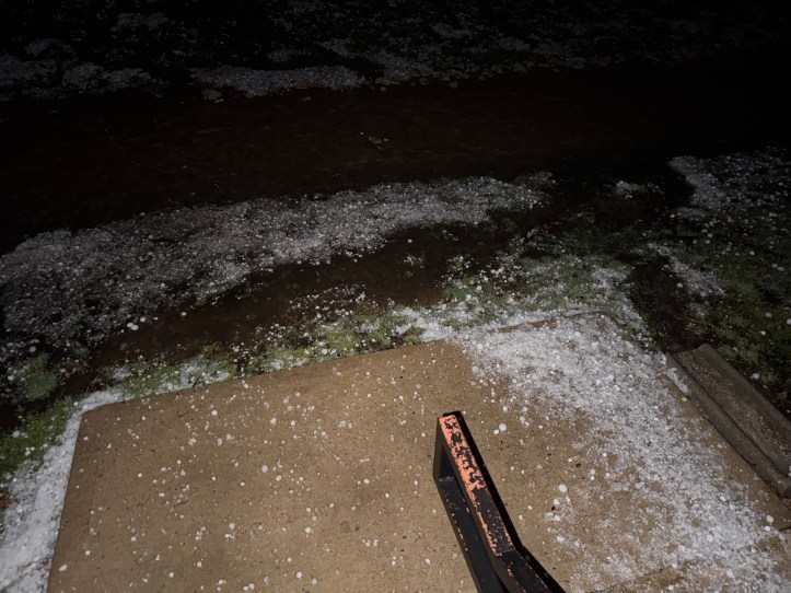 A nighttime scene showing a concrete step leading to a yard covered in scattered golf ball-sized hailstones. Some areas have accumulated small piles of hail, while the grass and pavement appear wet, reflecting light from an unseen source.