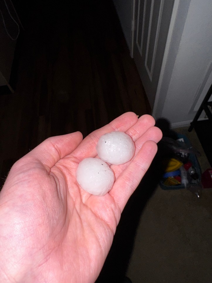 A close-up of a person’s hand holding two golf ball-sized hailstones indoors. The hailstones are smooth, icy, and slightly translucent, with small bits of debris embedded in them. The background is dimly lit, showing a hallway and household items.