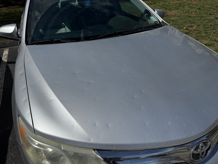 A silver Toyota Camry’s hood is covered in multiple small dents, likely from hail damage. Sunlight reflects off the metallic surface, emphasizing the dents. The windshield has water droplets on it, and the car is parked near a grassy area.