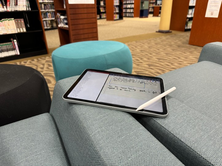 An iPad with an Apple Pencil rests on a light blue cushioned chair in a library. The screen displays a note-taking app with handwritten notes, suggesting it’s being used for studying or homework. In the background, bookshelves filled with books and manga, along with colorful ottomans, create a cozy study environment. The setting is quiet and inviting, ideal for focused work.
