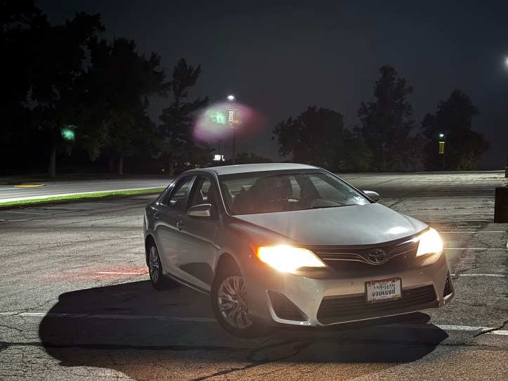 A silver Toyota Camry is parked in a dimly lit lot at night with its headlights on. The bright lights create lens flares in shades of green and pink. The surrounding area has streetlights, trees, and a few distant building lights.
